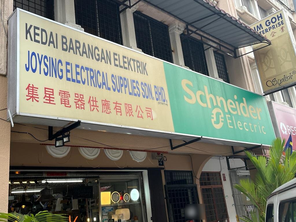 Shop signboard reading Joysing Electrical Supplies Sdn Bhd with a green Schneider Electric panel above a shopfront window