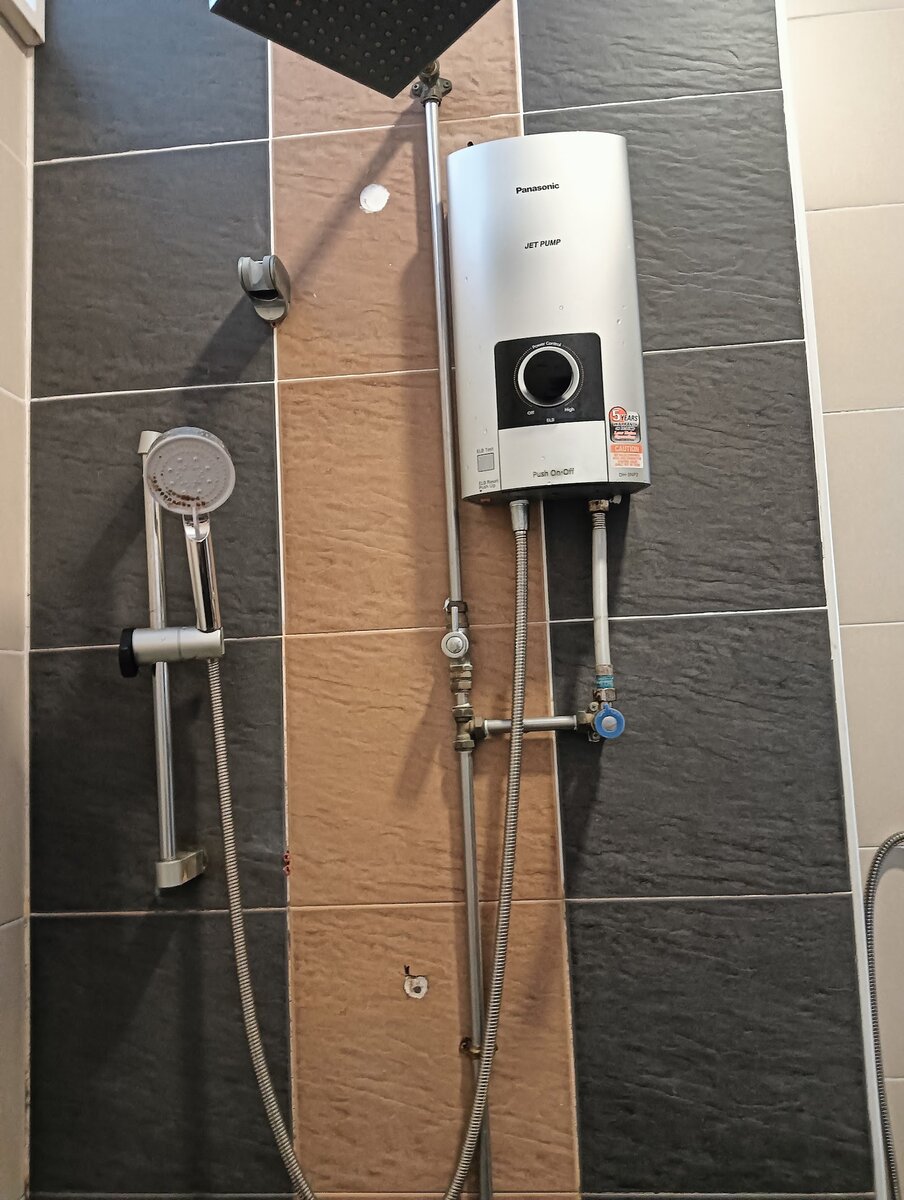 Silver Panasonic instant water heater mounted on dark and brown bathroom tiles, with a rain shower head above and a hand shower rail beside it