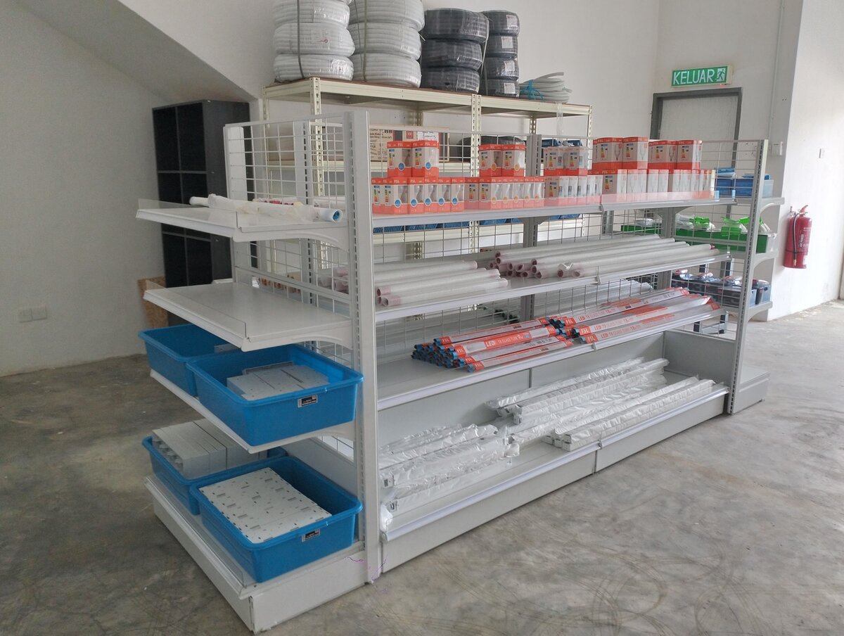 Shop aisle with shelves of orange-and-white FSL LED boxes, red-labelled LED tubes, white PVC conduit lengths, blue baskets and stacked conduit rolls on a rack behind