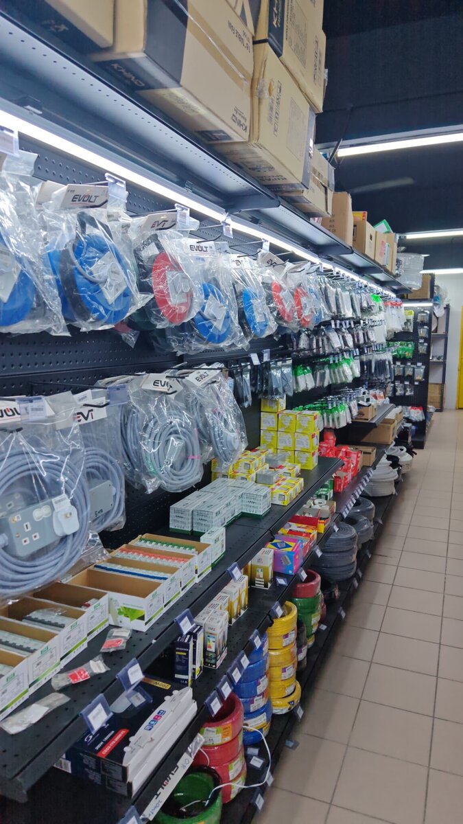 Long shop aisle with cable reels and extension leads hanging on pegboard, boxed switches and sockets, LED bulbs, a floodlight box and coils of coloured wire on the lower shelves