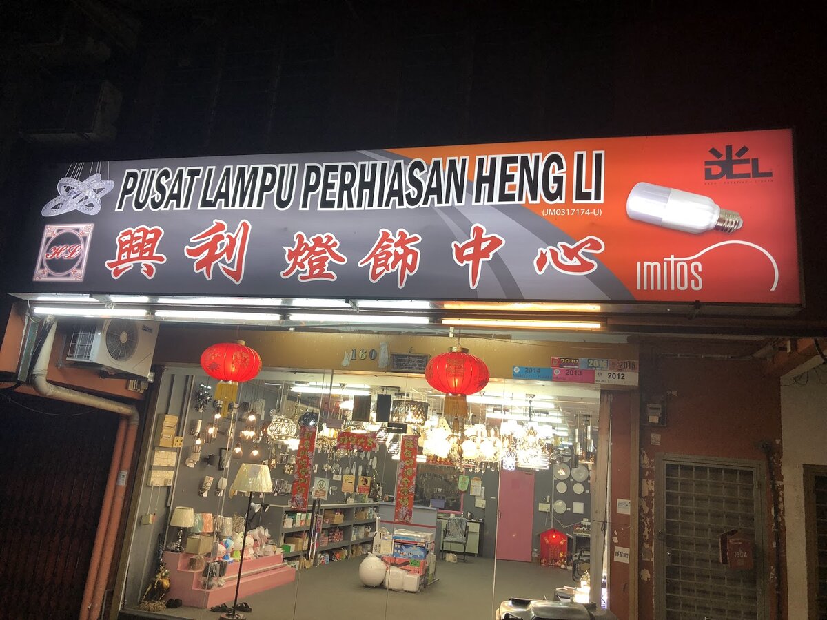 Lit red and grey signboard reading "Pusat Lampu Perhiasan Heng Li 興利燈飾中心" over a glass shopfront full of lamps and chandeliers