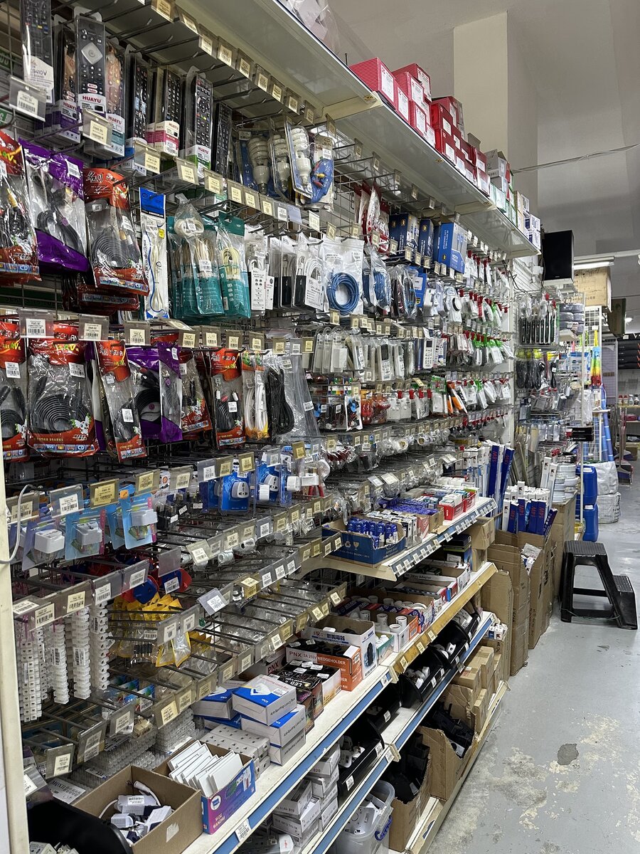 Long pegboard wall of packaged electrical accessories with price labels above shelves of boxed plugs, lamp holders and adaptors
