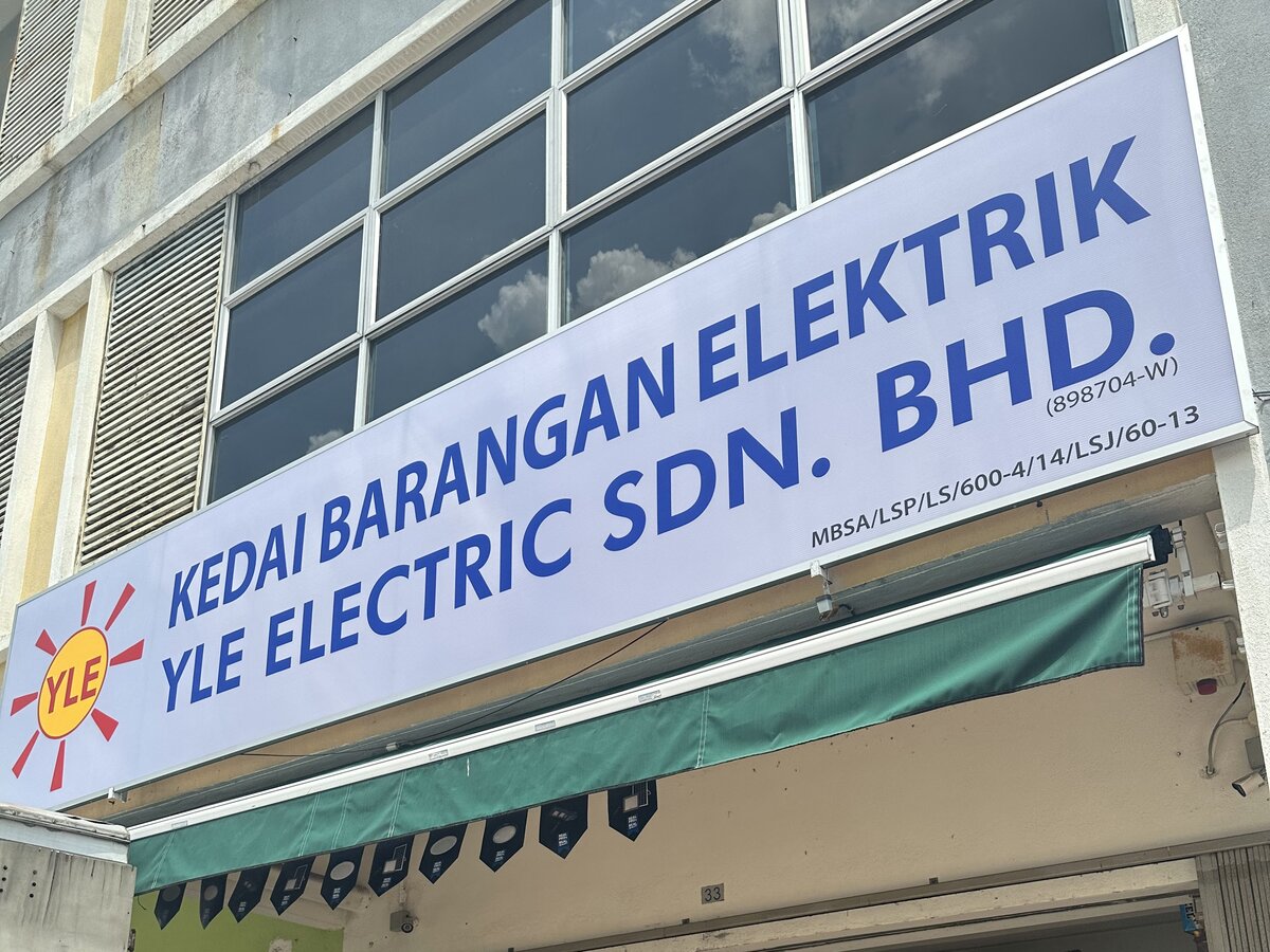 White shop signboard with blue lettering reading Kedai Barangan Elektrik, YLE Electric Sdn. Bhd., with a red and yellow sun logo, above a green awning
