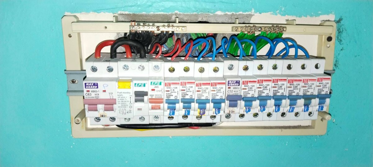 Cream DB box set into a turquoise wall with its cover off: a Maxguard C63 breaker, an RCCB marked 40A 100mA, then about a dozen MCBs of several makes, red, black, blue and green cables looping up to two brass terminal bars