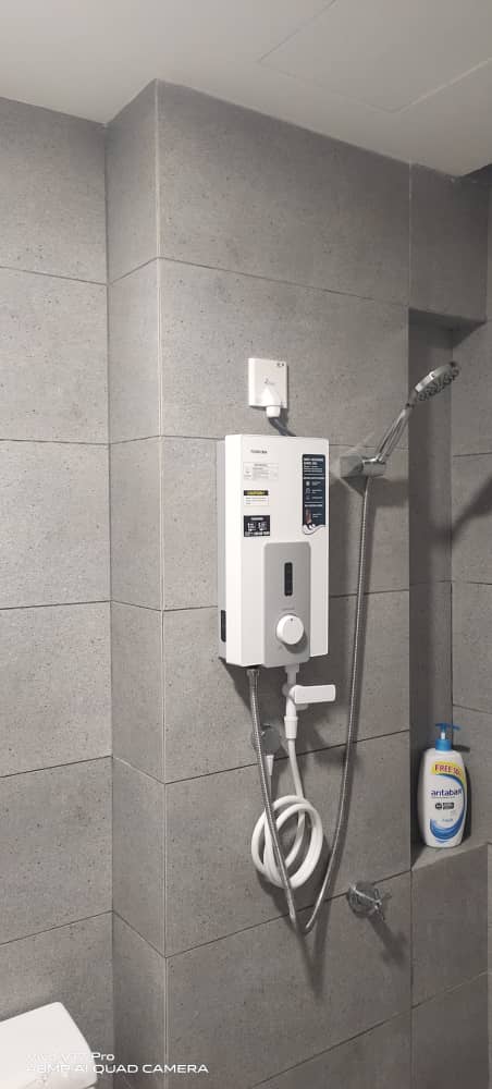 White instant water heater with a hand shower on a grey tiled bathroom wall, with a switch above it