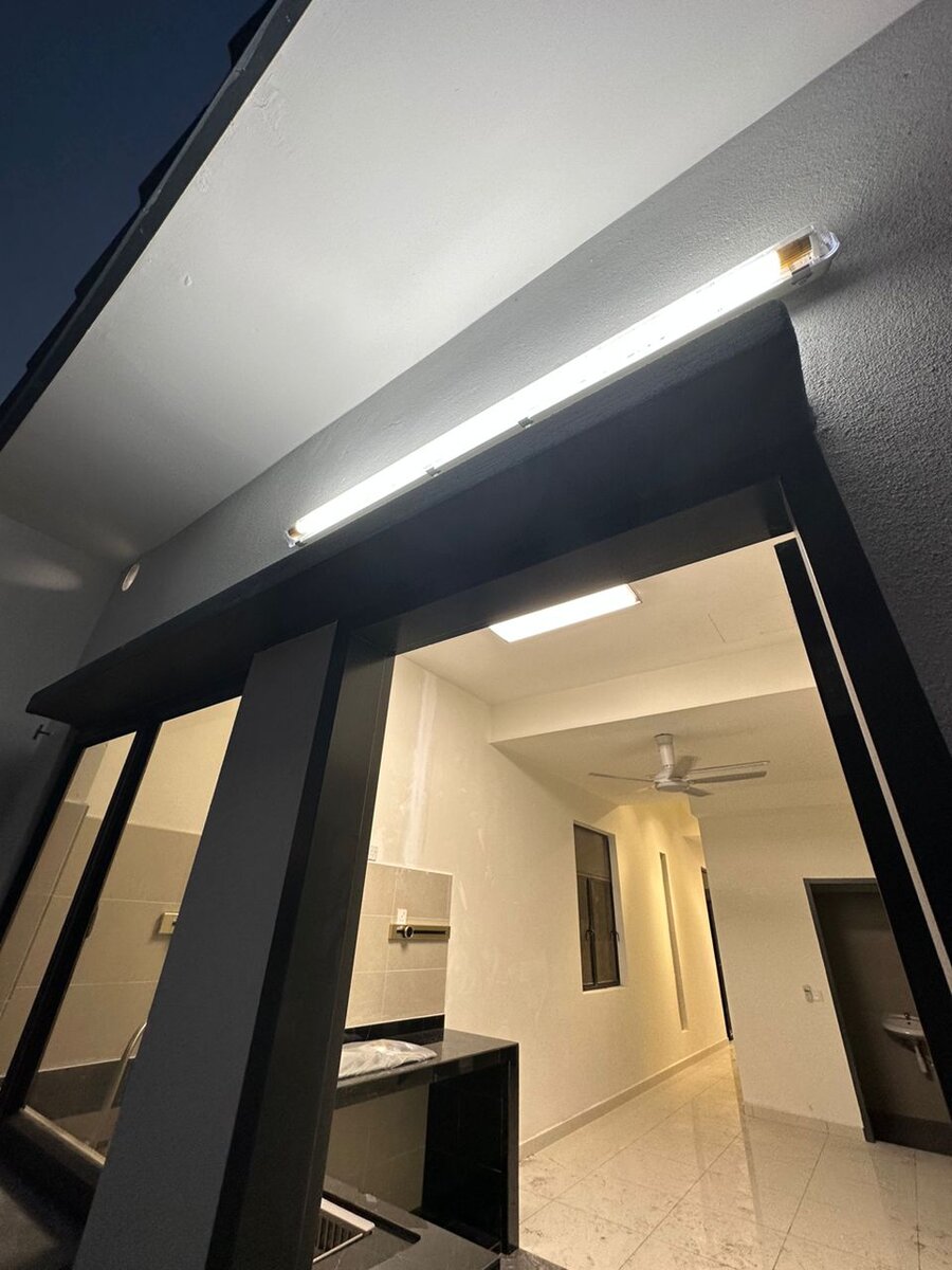 Night view of a long fluorescent batten lit under a roof overhang at the back of a house, looking through an opening into a lit kitchen and hallway with a ceiling fan