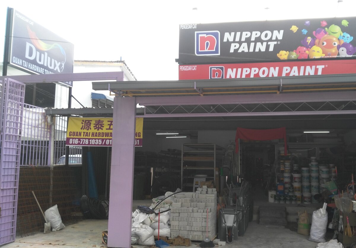 Open hardware shopfront under a purple steel canopy with Nippon Paint signboards above, a Dulux sign and a yellow banner reading Guan Tai Hardware with a phone number, and paint tins, bricks and sacks inside