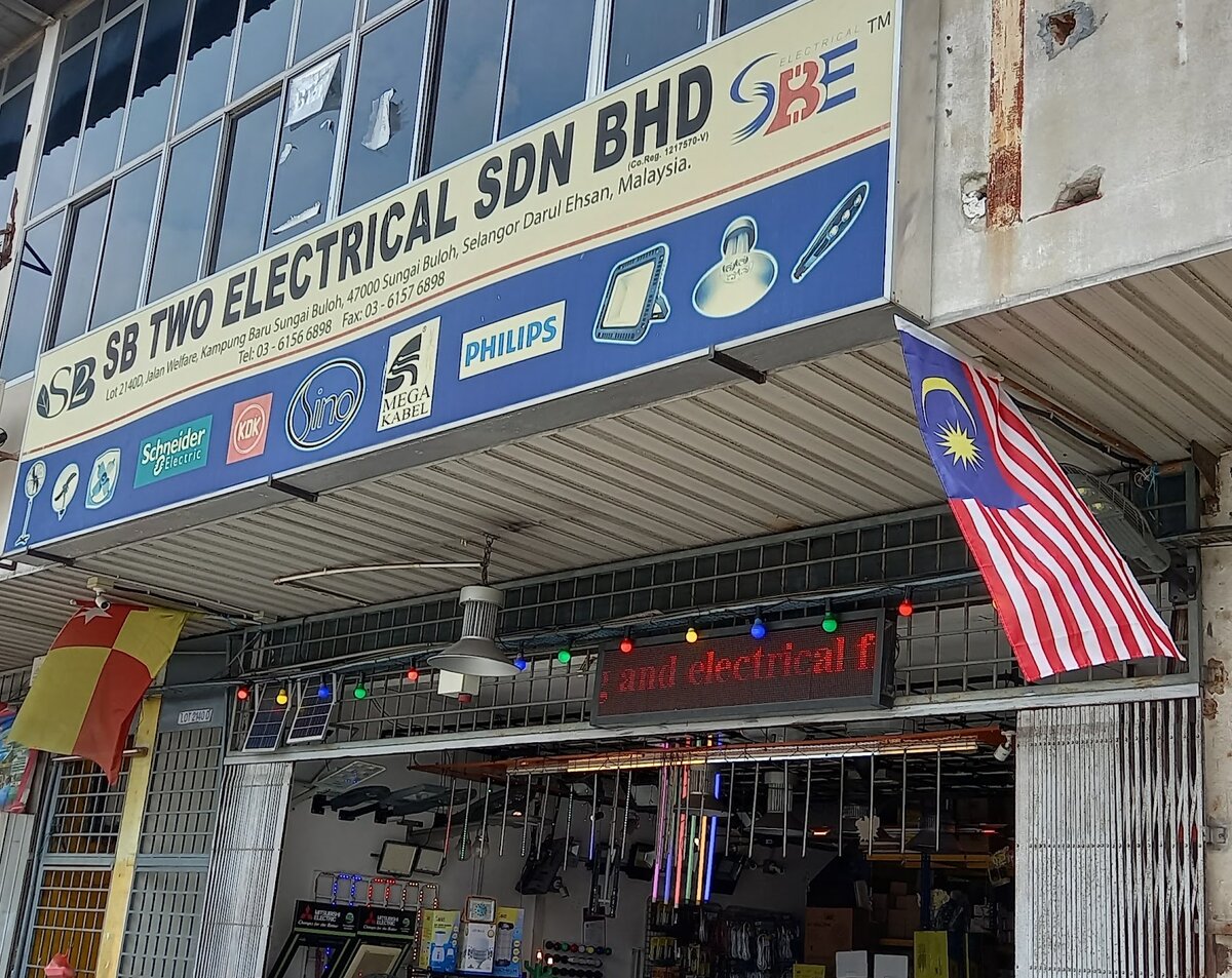 Shophouse with a cream and blue signboard reading SB Two Electrical Sdn Bhd, brand logos, a scrolling red LED sign and a Malaysian flag