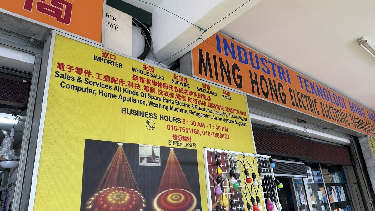 Yellow signboard with Chinese and English text beside an orange Ming Hong Electric Electronic Technologies sign, with coloured bulbs hanging below