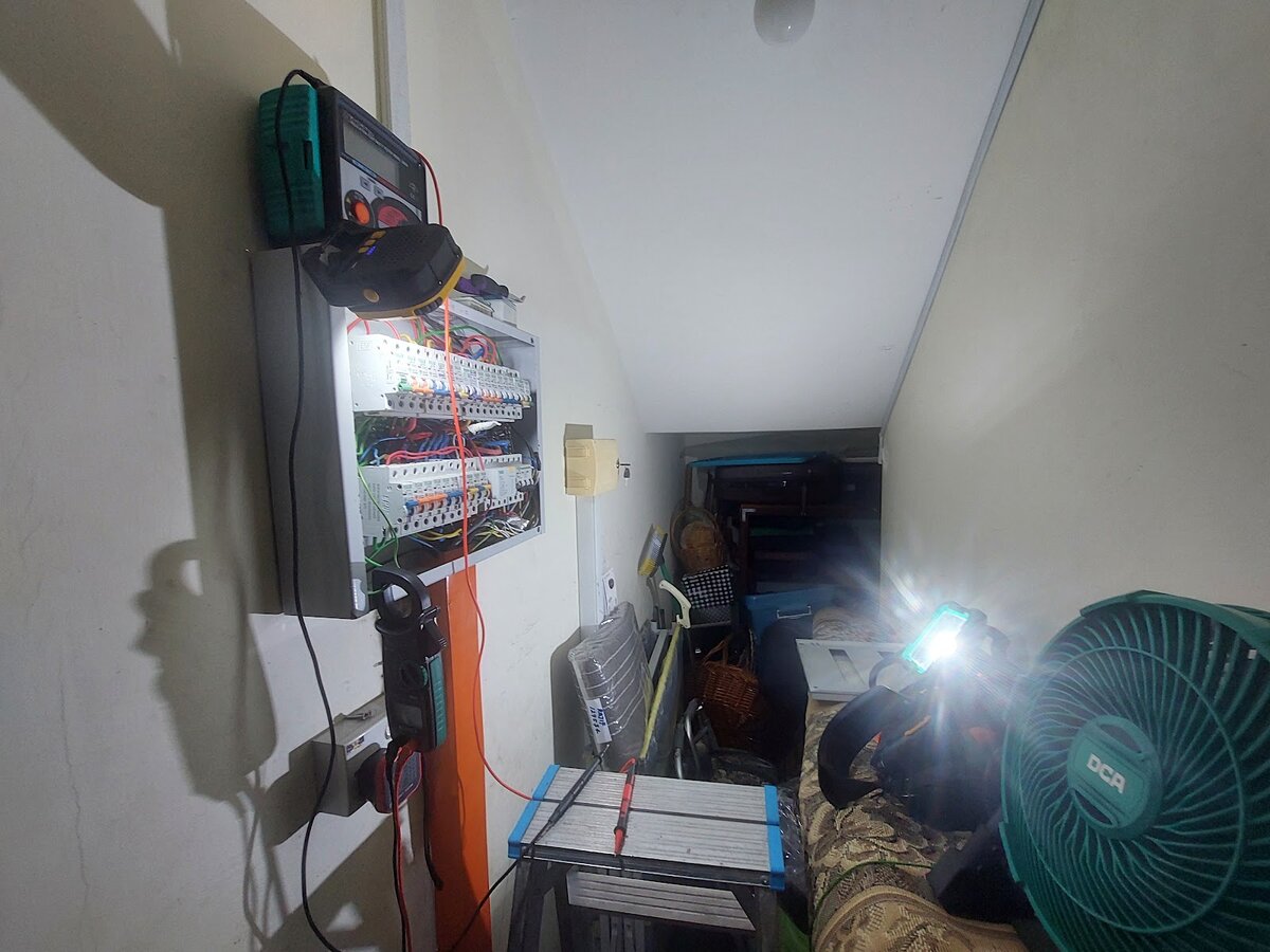 A distribution board on a wall in a cramped under-stair space, a tester on top of it and a clamp meter hanging below, a bright portable work light, a stool and a green fan in the foreground