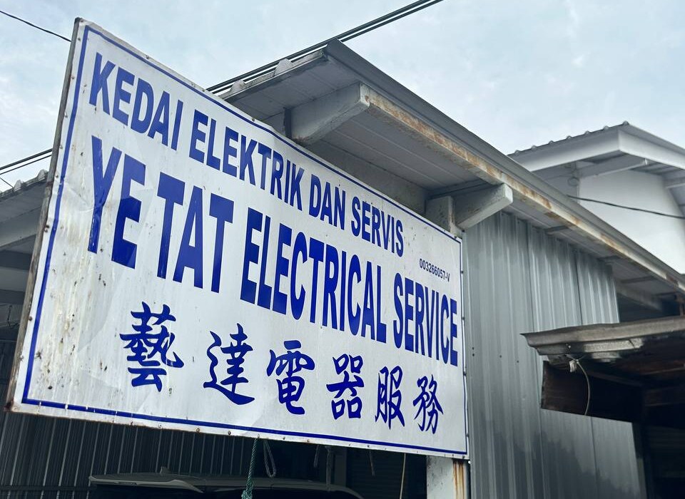 White signboard with blue lettering reading Kedai Elektrik dan Servis, Ye Tat Electrical Service and Chinese characters, mounted in front of a zinc-roofed building