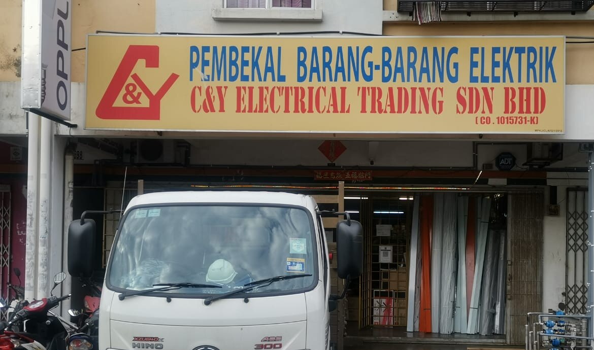 Yellow shop signboard with a red C&Y logo reading Pembekal Barang-Barang Elektrik, C&Y Electrical Trading Sdn Bhd, above an open shopfront with a white lorry parked in front