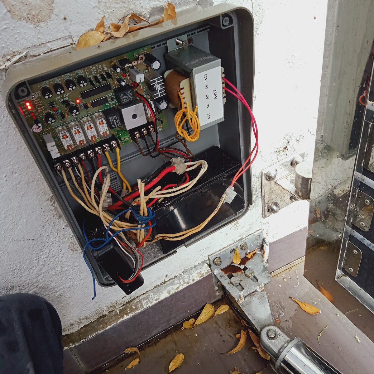 Open auto gate control box with a circuit board and wiring, mounted on a wall beside a gate arm