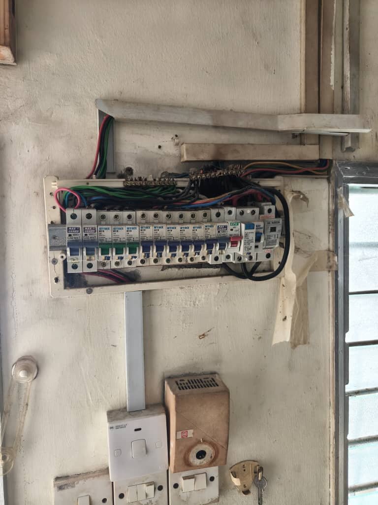 Worn wall-mounted board with a row of old miniature circuit breakers, loose cables above it and taped edges, switches and a doorbell transformer below