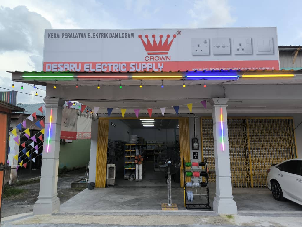 Single-storey shop with a white signboard reading Desaru Electric Supply and a red Crown logo beside pictures of wall switches and sockets