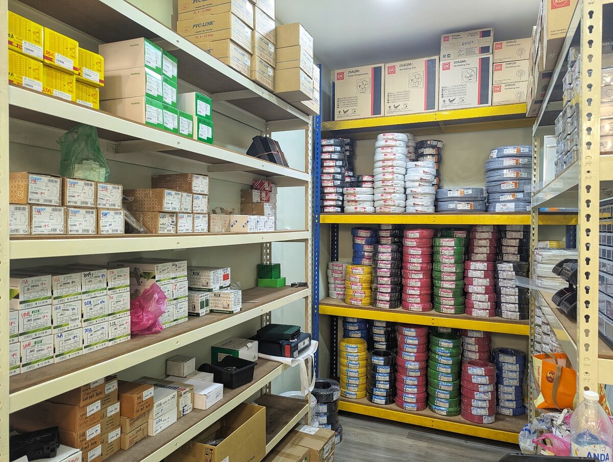 Store room with shelves of boxed circuit breakers and consumer units on the left and yellow racks of red, black, green, blue and yellow cable coils under KDK fan boxes on the right