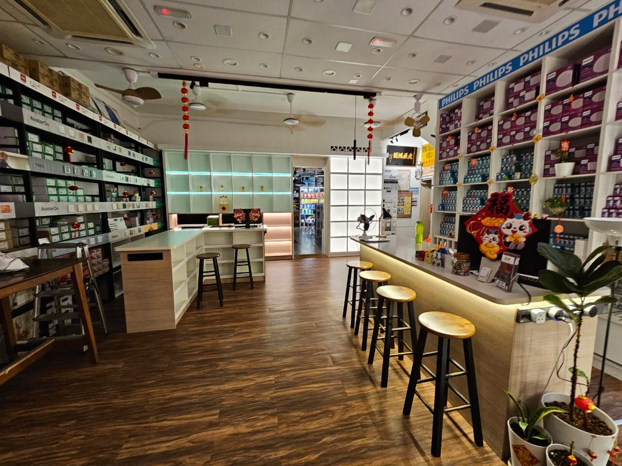 Shop interior with wooden floor, shelves of boxed switches and switch-plate samples on the left, a wall of purple and blue Philips lamp boxes on the right and a service counter with stools