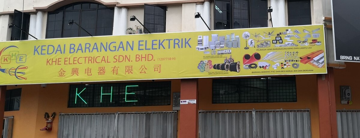 Long yellow shop signboard reading Kedai Barangan Elektrik, KHE Electrical Sdn. Bhd. and Chinese characters, with product pictures of distribution boards, cable drums, fans, LED lights and PVC conduit, above closed shutters and a green neon KHE sign
