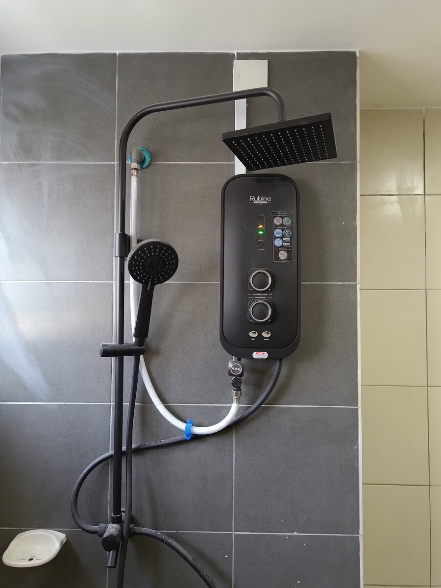 Black instant water heater with two dials and indicator lights mounted on a dark grey tiled wall below a square rain shower head, with a hand shower on a rail