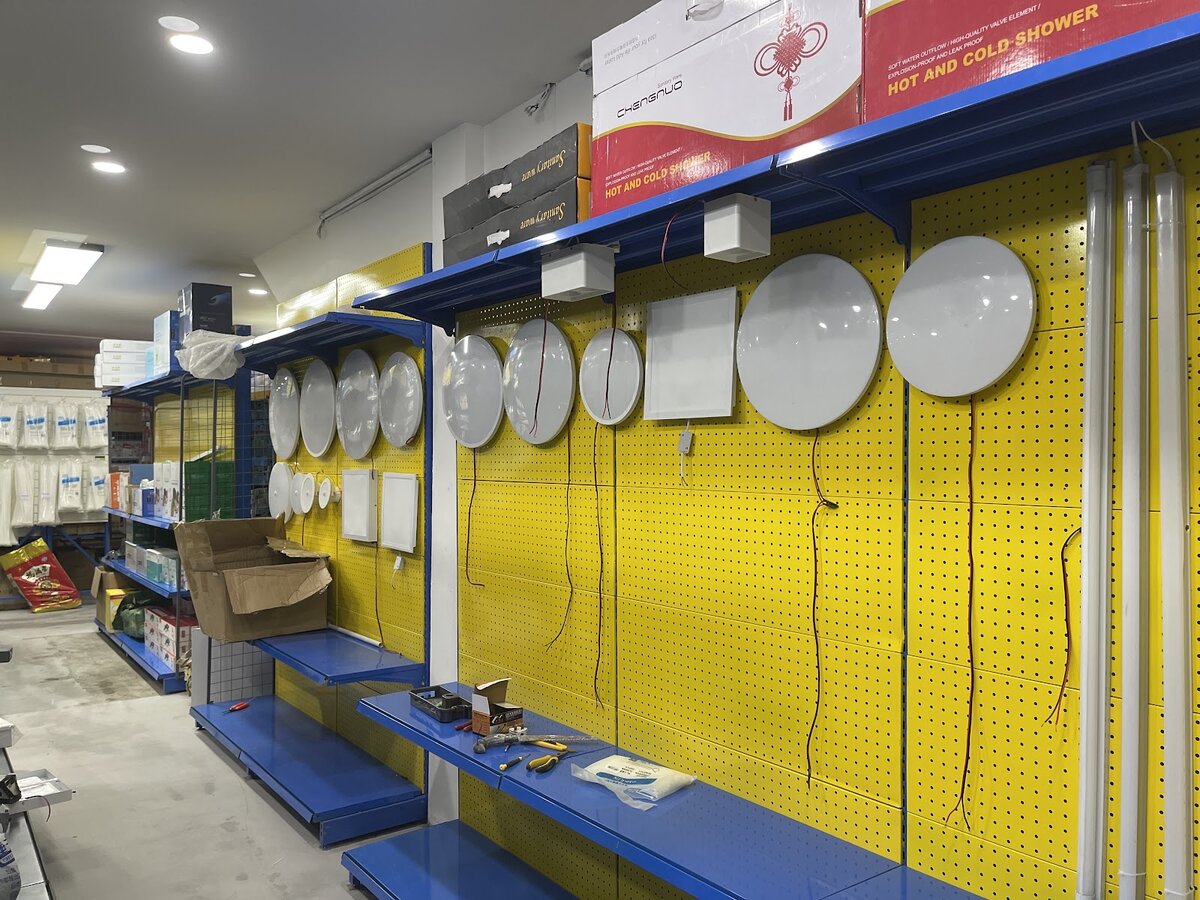 Yellow pegboard wall hung with round white LED ceiling lamps, square LED panels and long LED tubes, with boxed shower heaters on the shelf above