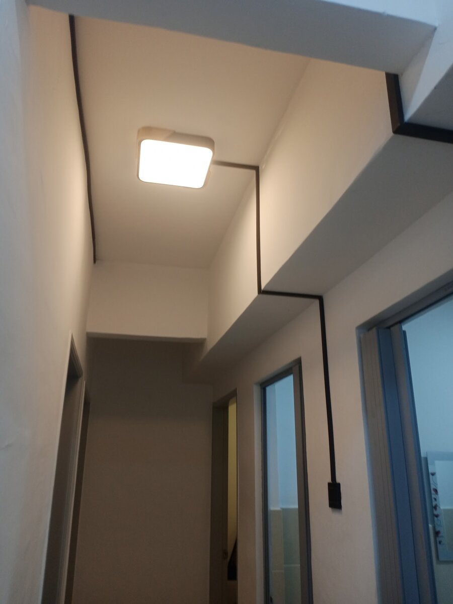Narrow corridor with a square ceiling lamp and a black conduit running along the ceiling and down the wall to a switch