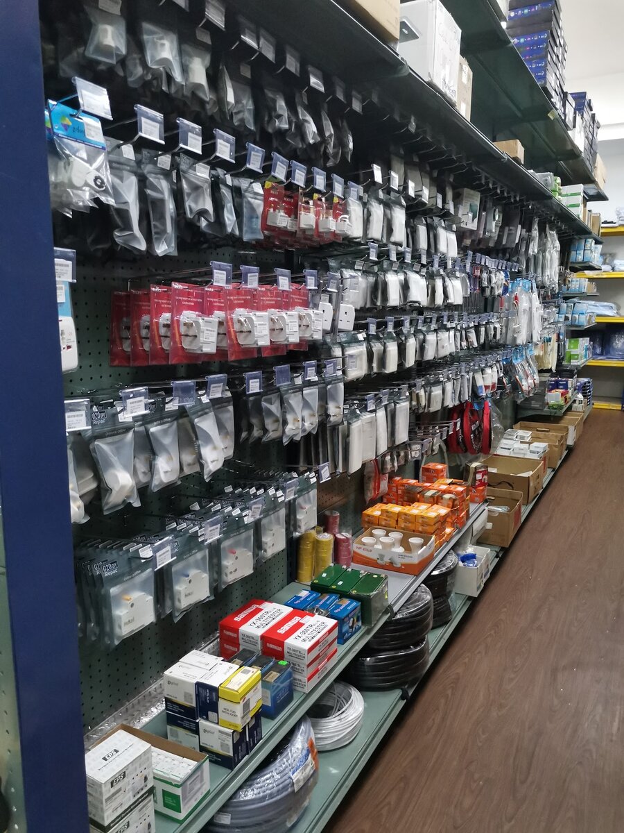 Pegboard aisle of packed plug tops, adaptors and switch sockets, with boxed multitesters, an RCCB box, lamp holders and coils of cable on the lower shelves