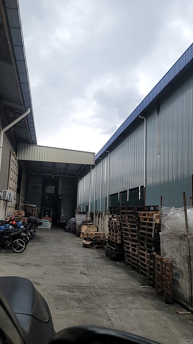 Concrete yard between two large metal-clad warehouse sheds, with stacked wooden pallets on the right and motorcycles parked on the left