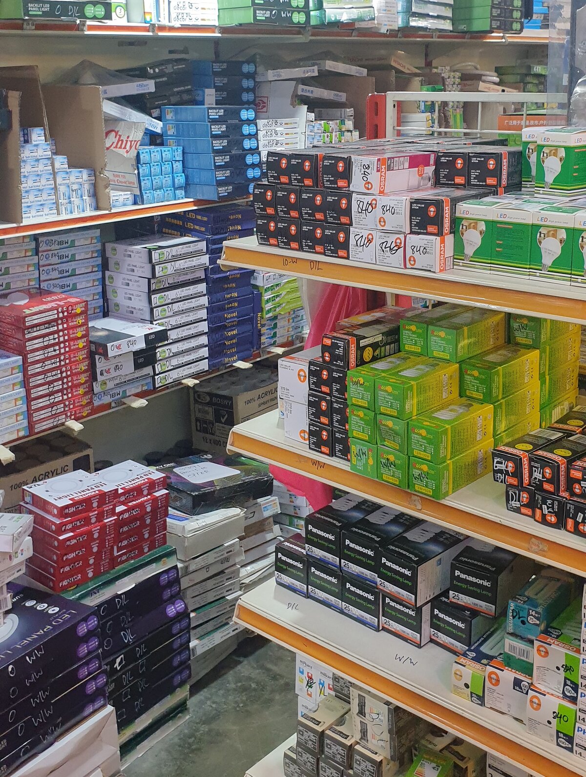 Shop shelves stacked with boxed lamps and LED panel lights: Philips boxes on the upper left, Osram LED Value Stick and Panasonic lamp boxes on the right-hand shelves