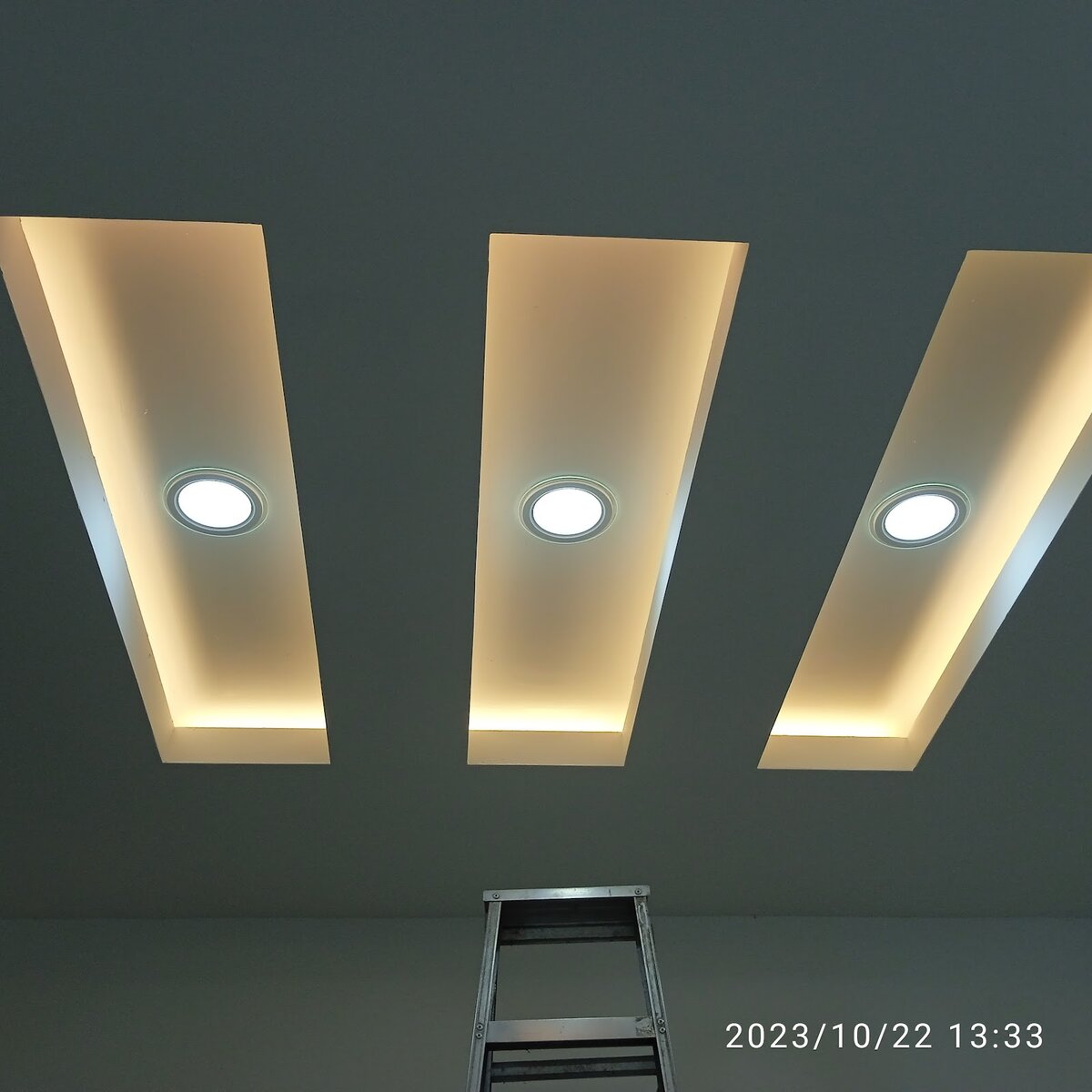 Three recessed ceiling panels with warm cove lighting and a round downlight in each, with a ladder below