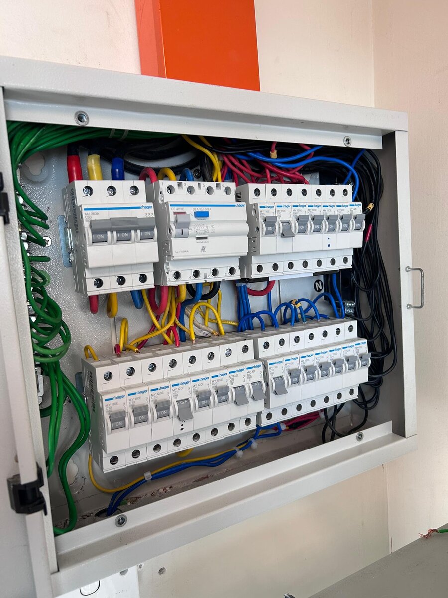 Open grey distribution board on a wall with white Hager breakers in two rows, red, yellow and blue cables at the top and green earth wires on the left
