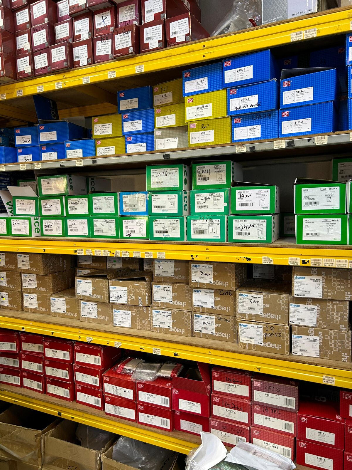 Yellow shelves stacked with maroon, blue, yellow, green, brown and red boxes of wall switches and sockets, with handwritten labels