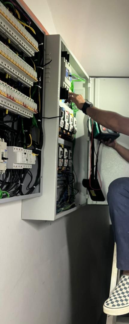 Tall grey distribution board with its door open, rows of miniature circuit breakers and moulded-case breakers inside, and a technician's arm holding a test meter lead to it