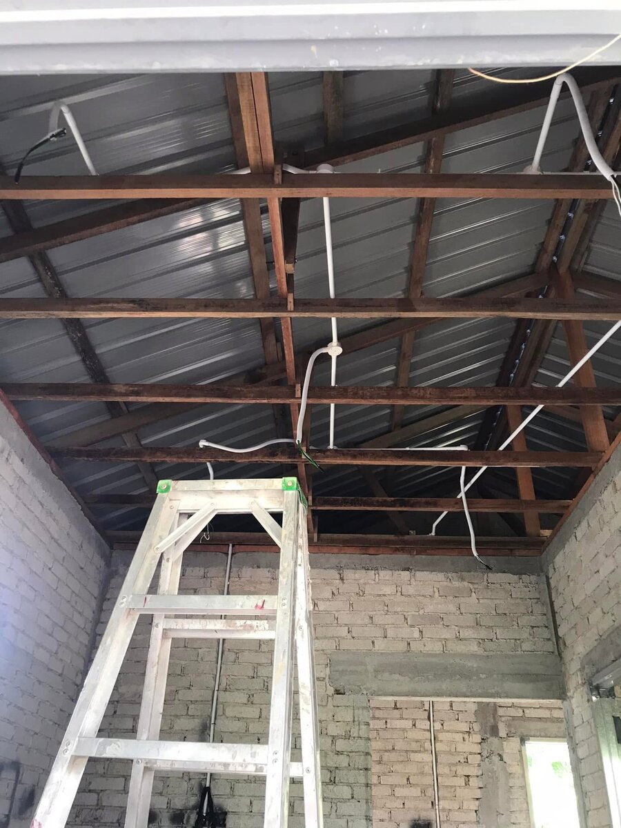 Unplastered brick room open to a metal roof, with white PVC conduits running along wooden trusses and cable ends hanging down, and an aluminium ladder in the foreground