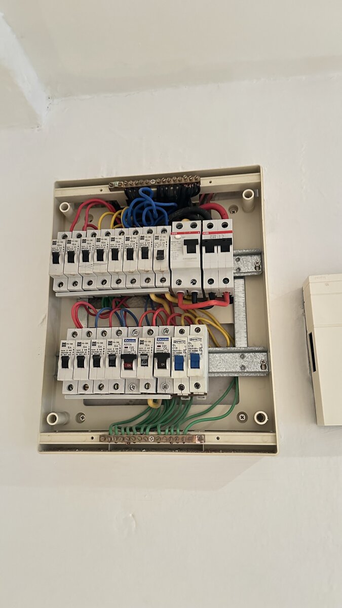 Open beige distribution board with two rows of white MCBs, red, yellow, blue and black cables, an earth bar at the bottom and two red-banded ABB devices at the top right