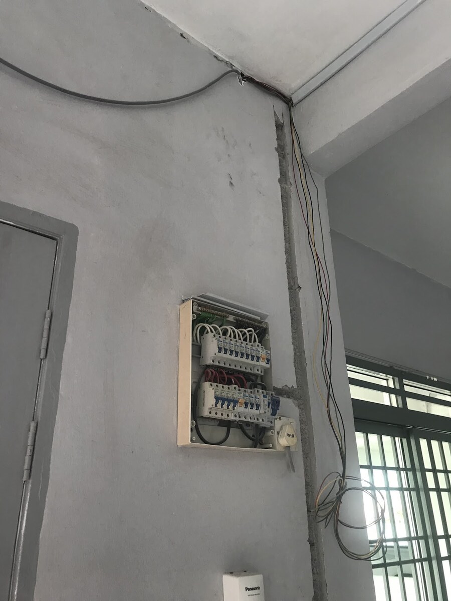 Open distribution board with two rows of breakers on a grey wall, a chased channel running up beside it to the ceiling and loose coloured wires hanging by a window grille