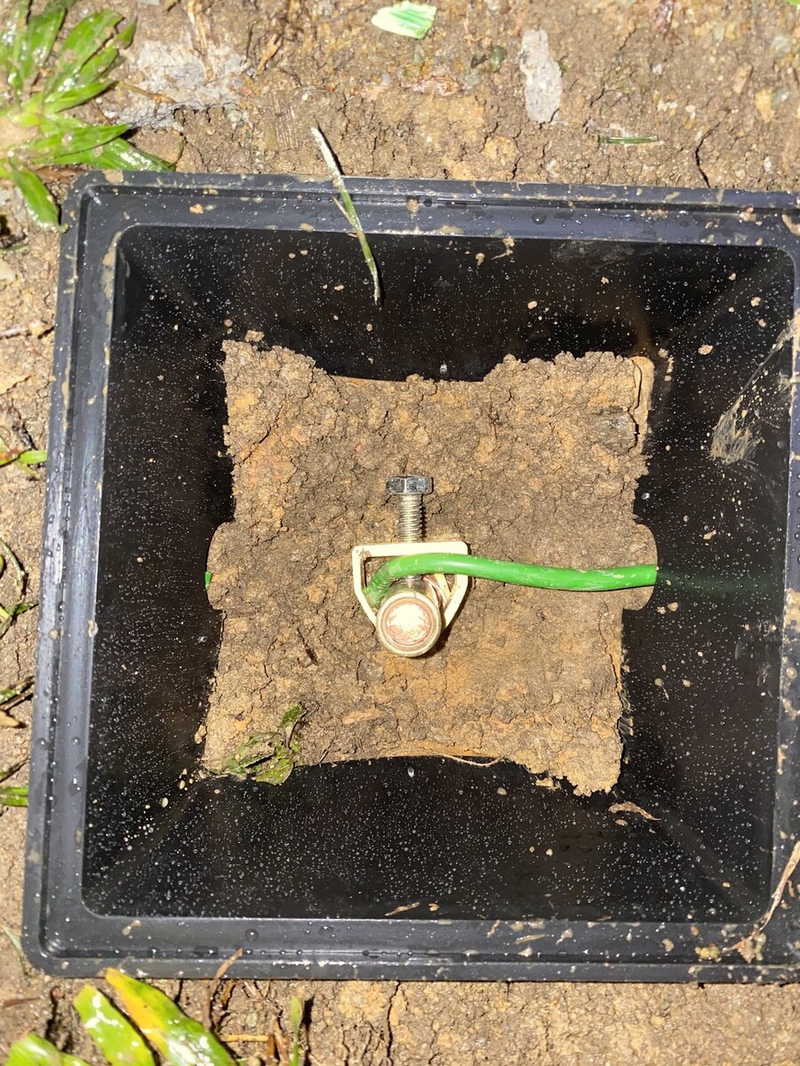 Black square earth pit set in soil with a copper earth rod clamp and a green earth cable