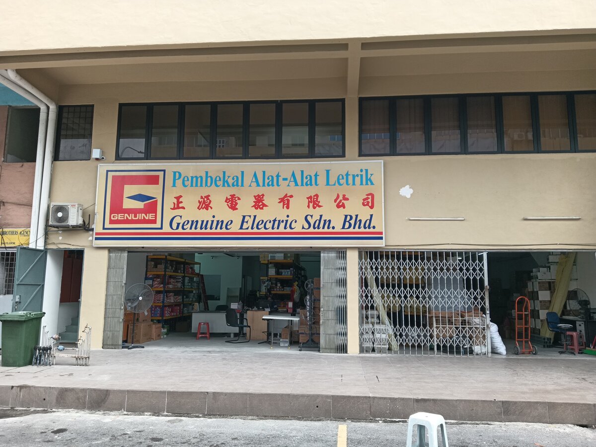 Two shop units in a cream-painted block, one open with a counter, fans and shelves of boxed electrical stock, the other partly behind a folding grille with cartons and conduit, under a signboard reading Pembekal Alat-Alat Letrik Genuine Electric Sdn Bhd