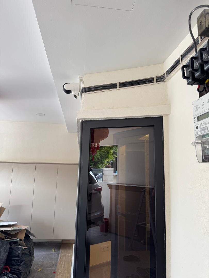 Corner of a house porch above a black-framed glass door, with a CCTV camera, black cables in an open white trunking along the top of the wall and a TNB meter mounted beside the door