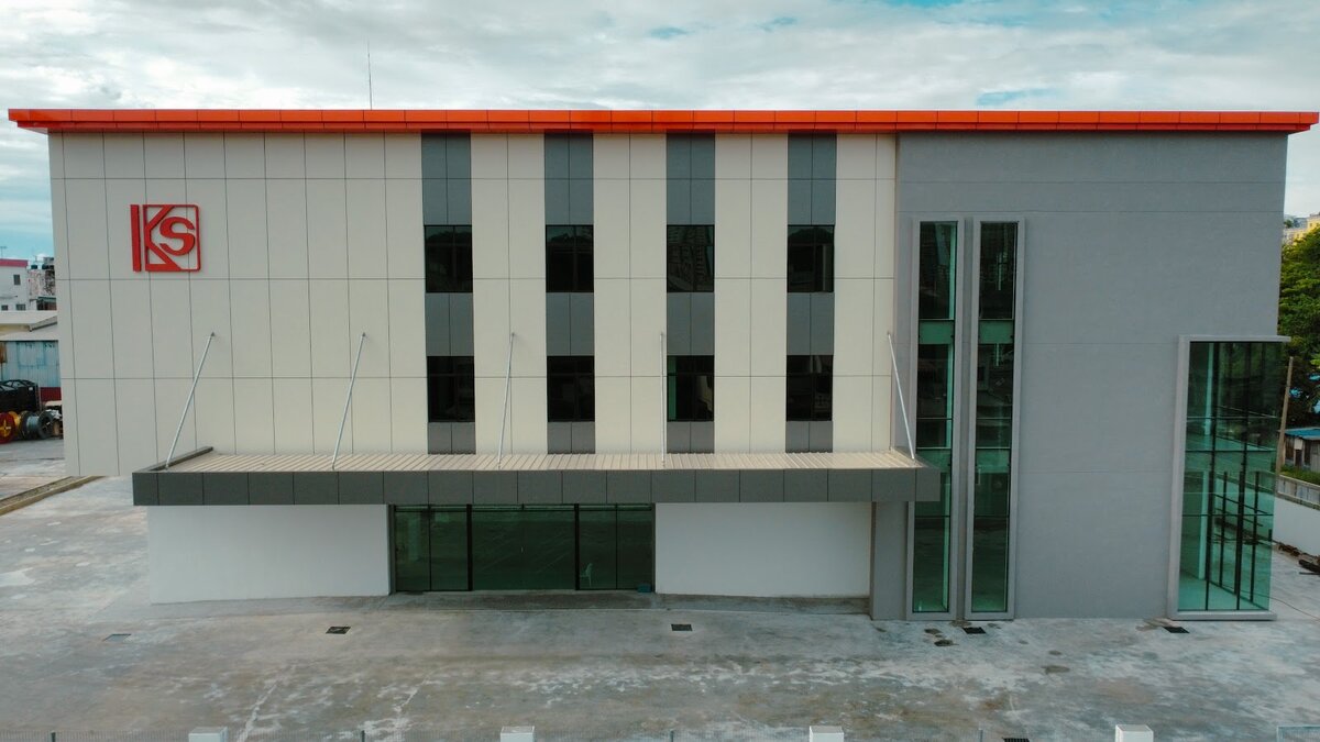 Grey and white three-storey commercial building with a red roof trim, KS logo, tall windows and a paved forecourt