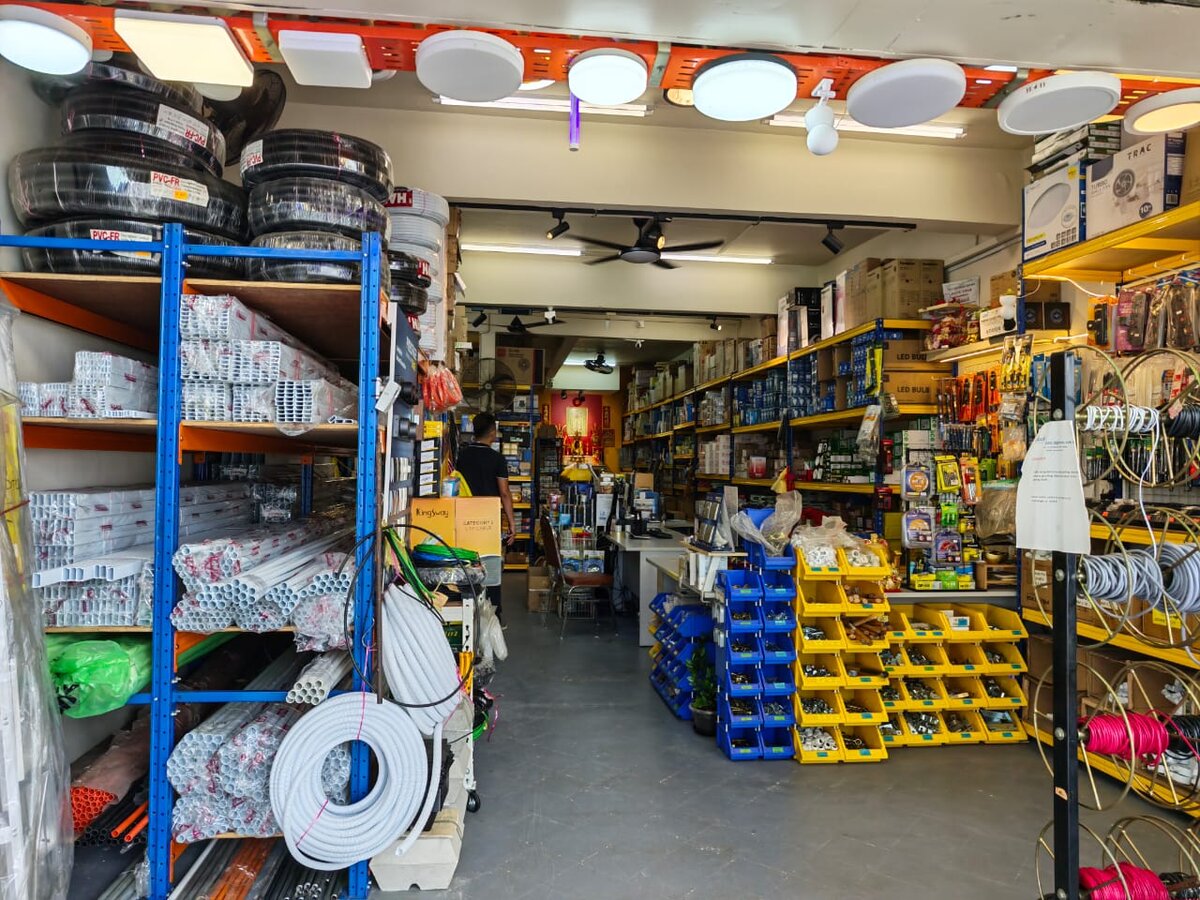 Electrical shop interior with blue racks of white PVC conduit and trunking, black flexible conduit coils, yellow and blue parts bins, cable reels and round LED ceiling lights overhead