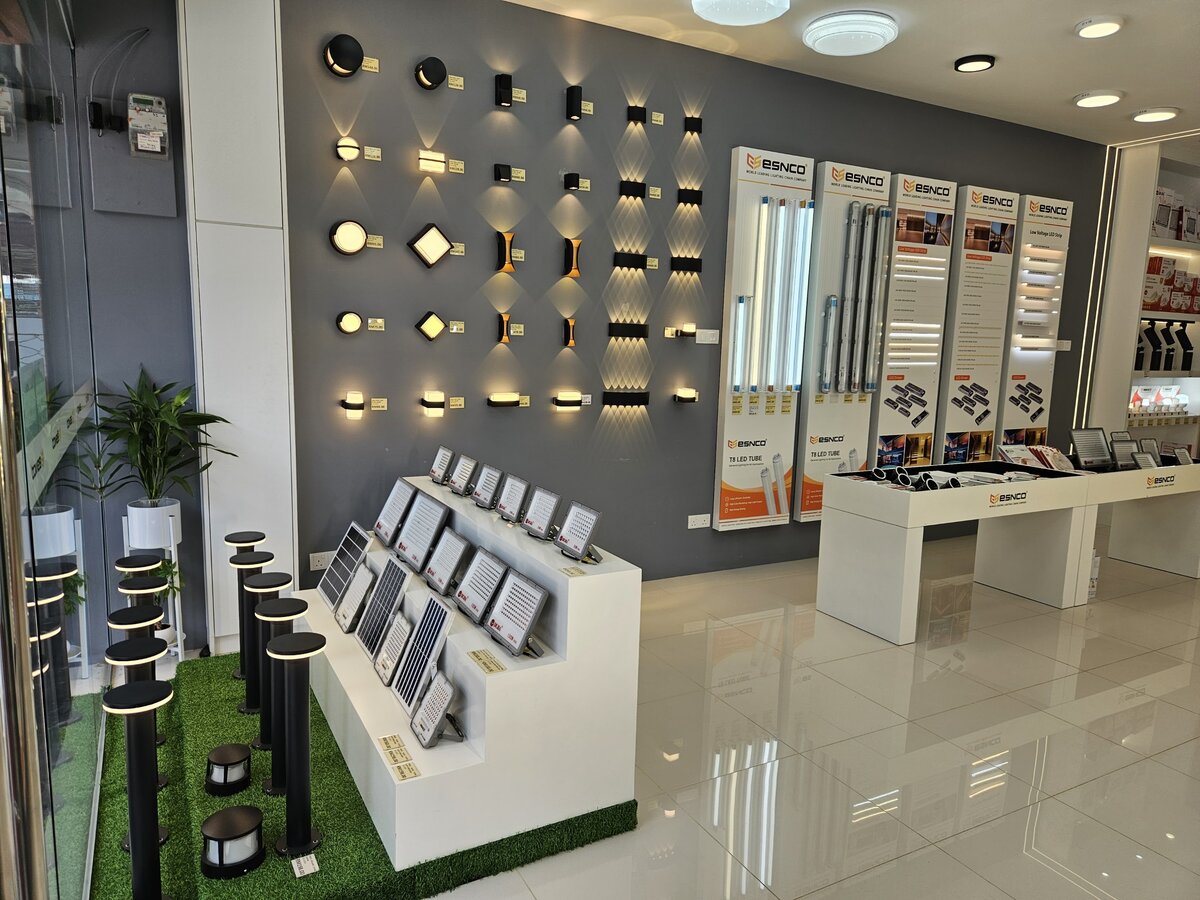 Grey showroom wall of lit black outdoor wall lights with yellow price tags, a tiered white stand of LED floodlights and solar floodlights, and tall orange-and-white Esnco display boards of LED tubes and strip lights
