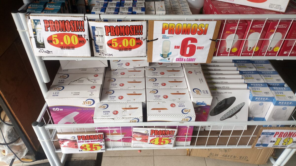 White wire rack of boxed lamps and UFO-style LED lamps with red and yellow Promosi price cards from RM5 to RM45