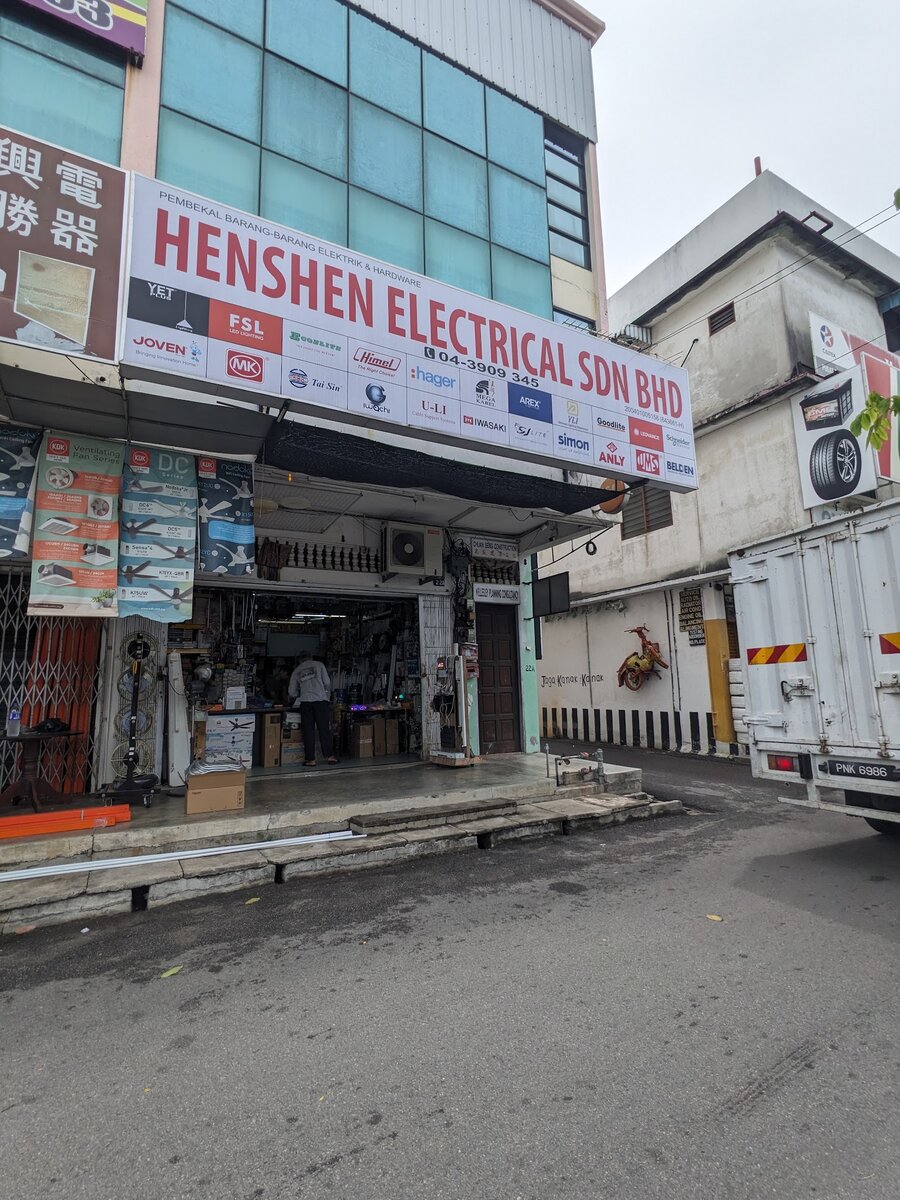 Corner shophouse with a white Henshen Electrical Sdn Bhd signboard covered in brand logos, an open shopfront with fans and boxes, and a lorry parked beside it