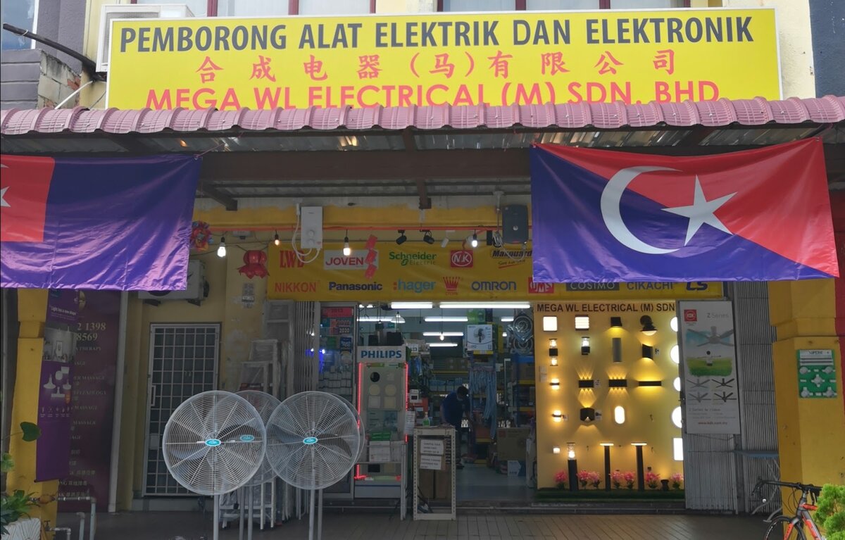 Shopfront with a yellow signboard reading Mega WL Electrical (M) Sdn. Bhd. in Malay, Chinese and English, a brand banner showing Schneider Electric, Hager, Maxguard and Omron, stand fans outside and lit wall lamps on display