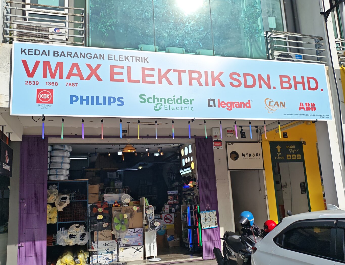 Shop under a light-blue signboard reading Kedai Barangan Elektrik, VMax Elektrik Sdn. Bhd., with brand logos, white conduit coils and shelves of trunking on the left and boxed ceiling fans at the door