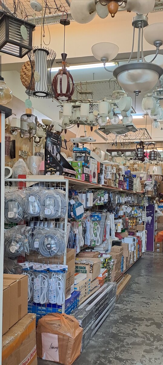 Narrow shop aisle under dozens of hanging pendant lamps and chandeliers, with wire racks of grey extension cords and shelves of boxed goods