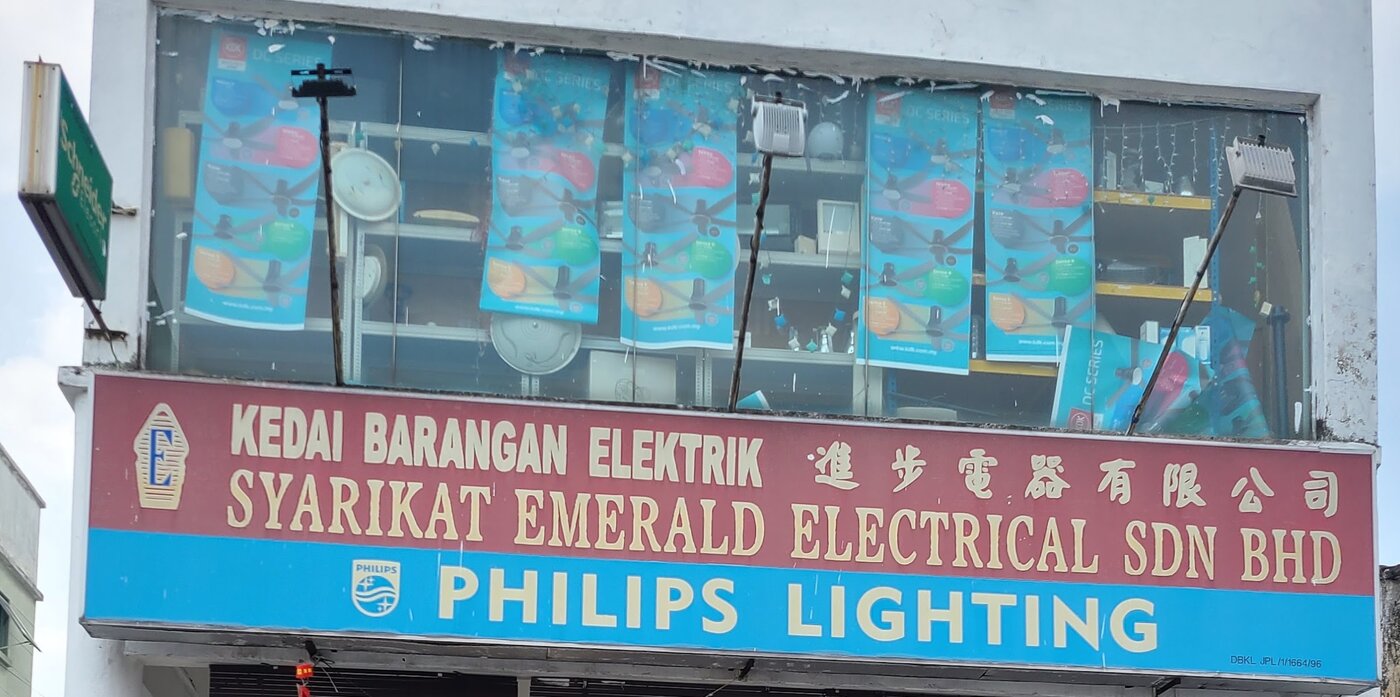Maroon and blue fascia reading Syarikat Emerald Electrical Sdn Bhd and Philips Lighting, below a window with ceiling-fan posters and a green Schneider sign at the corner
