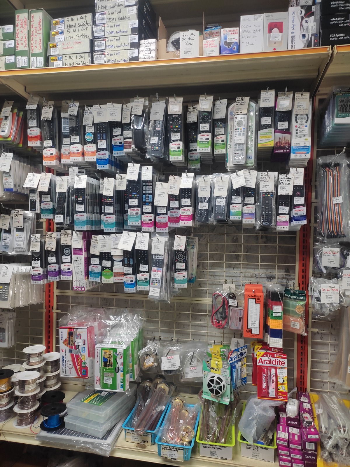 Pegboard wall of packaged remote controls with HDMI switch boxes on a shelf above, and spools of wire, breadboards, heating elements and glue packs on the shelf below