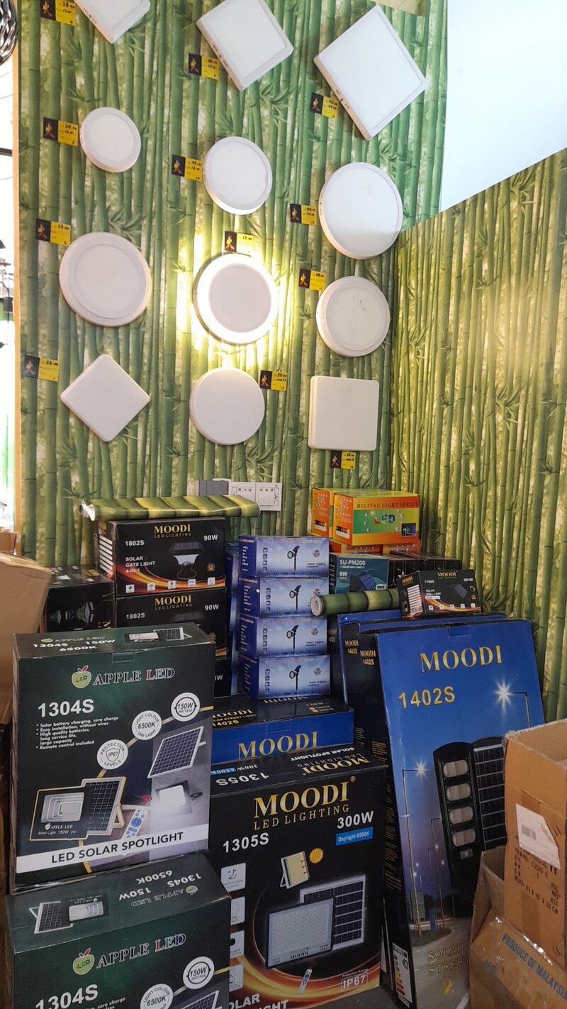 Wall of round and square LED ceiling panels above stacked boxes of Moodi and Apple LED solar lights