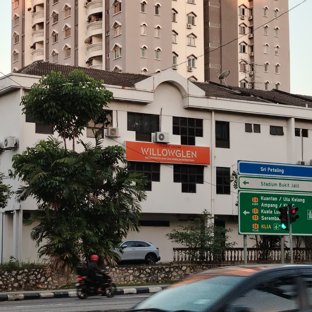 White low-rise office building with an orange Willowglen sign, flats behind and road signs for Sri Petaling in front